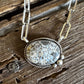 A silver necklace with a textured oval pendant featuring a mosaic of light blue and white pieces, resting on a rustic, weathered wooden surface.