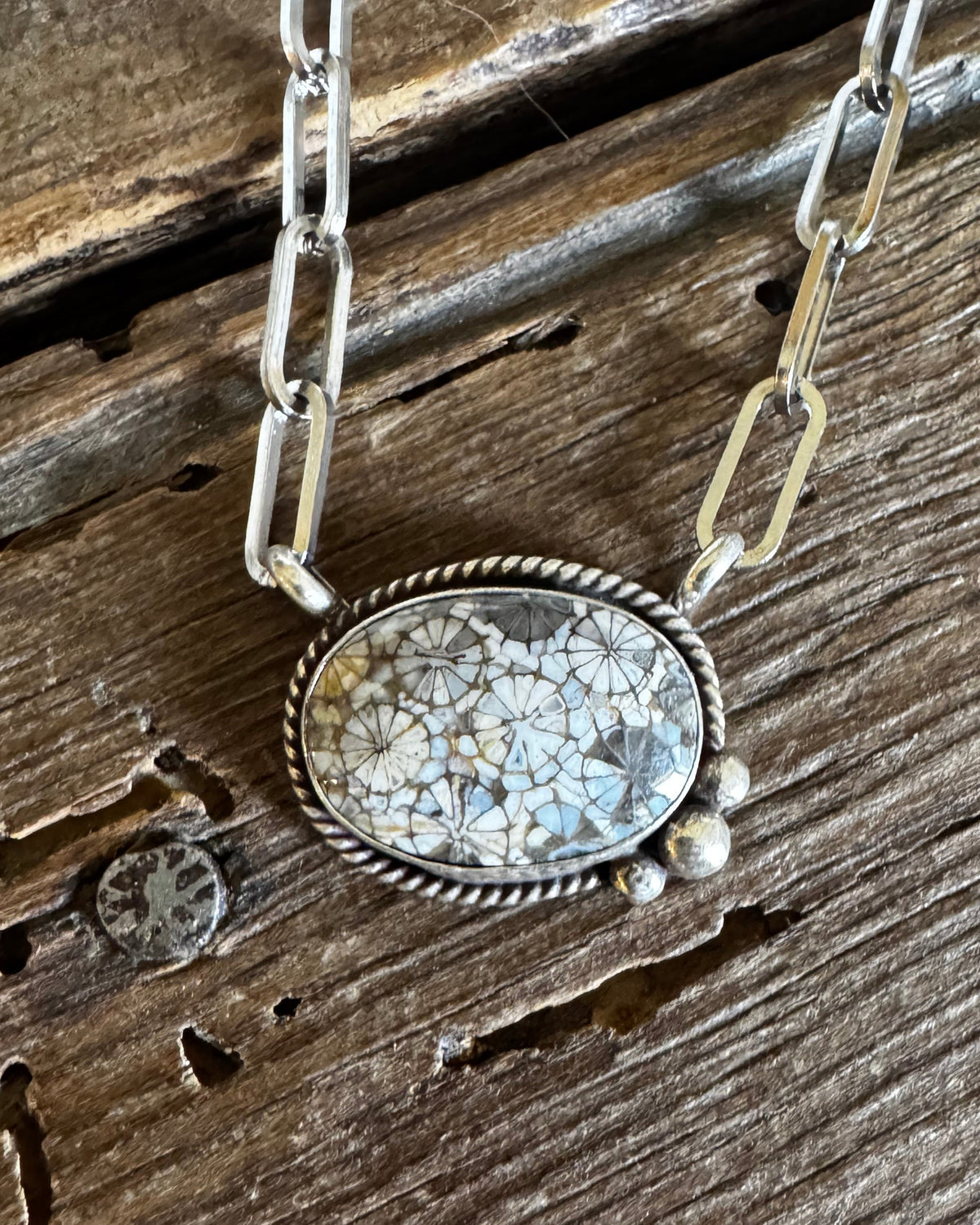 A silver necklace with a textured oval pendant featuring a mosaic of light blue and white pieces, resting on a rustic, weathered wooden surface.