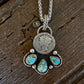 A silver pendant necklace featuring a circular coin charm and a paw print design set with turquoise stones, displayed on a rustic dark wood surface.