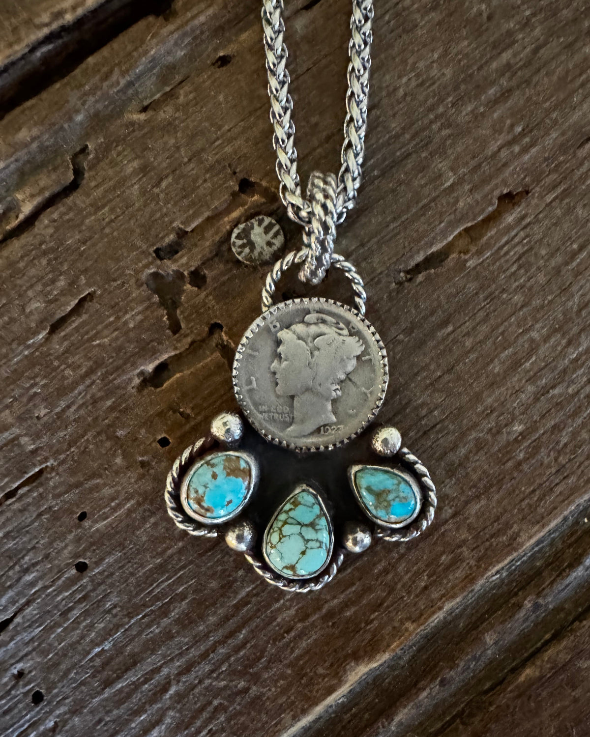 A silver pendant necklace featuring a circular coin charm and a paw print design set with turquoise stones, displayed on a rustic dark wood surface.