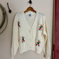 White cardigan with embroidered designs on a hanger against a plain wall.
