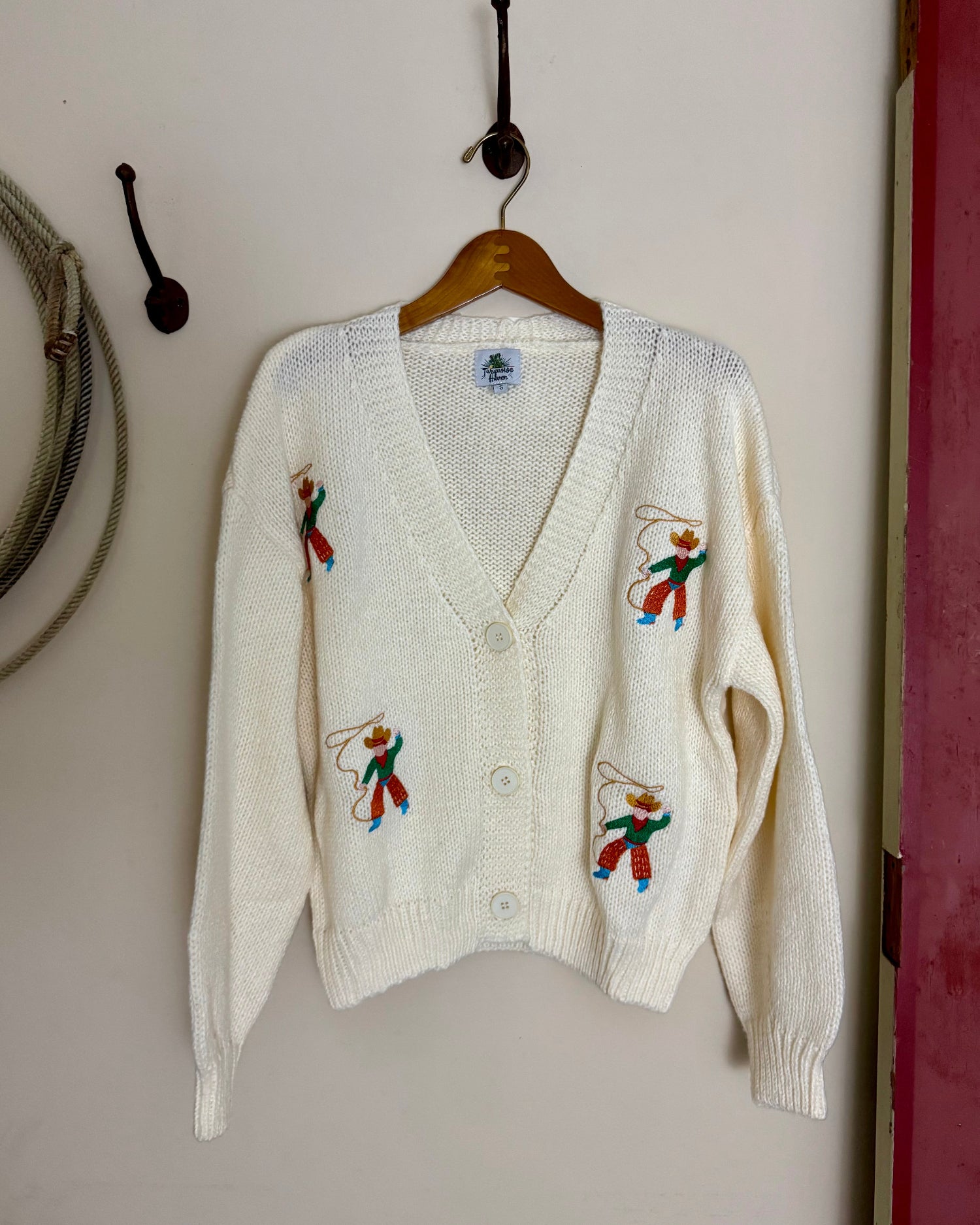 White cardigan with embroidered designs on a hanger against a plain wall.