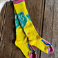 A pair of bright yellow crew socks with pink, teal, and blue graphic details, laid flat on a rustic dark wood plank surface.