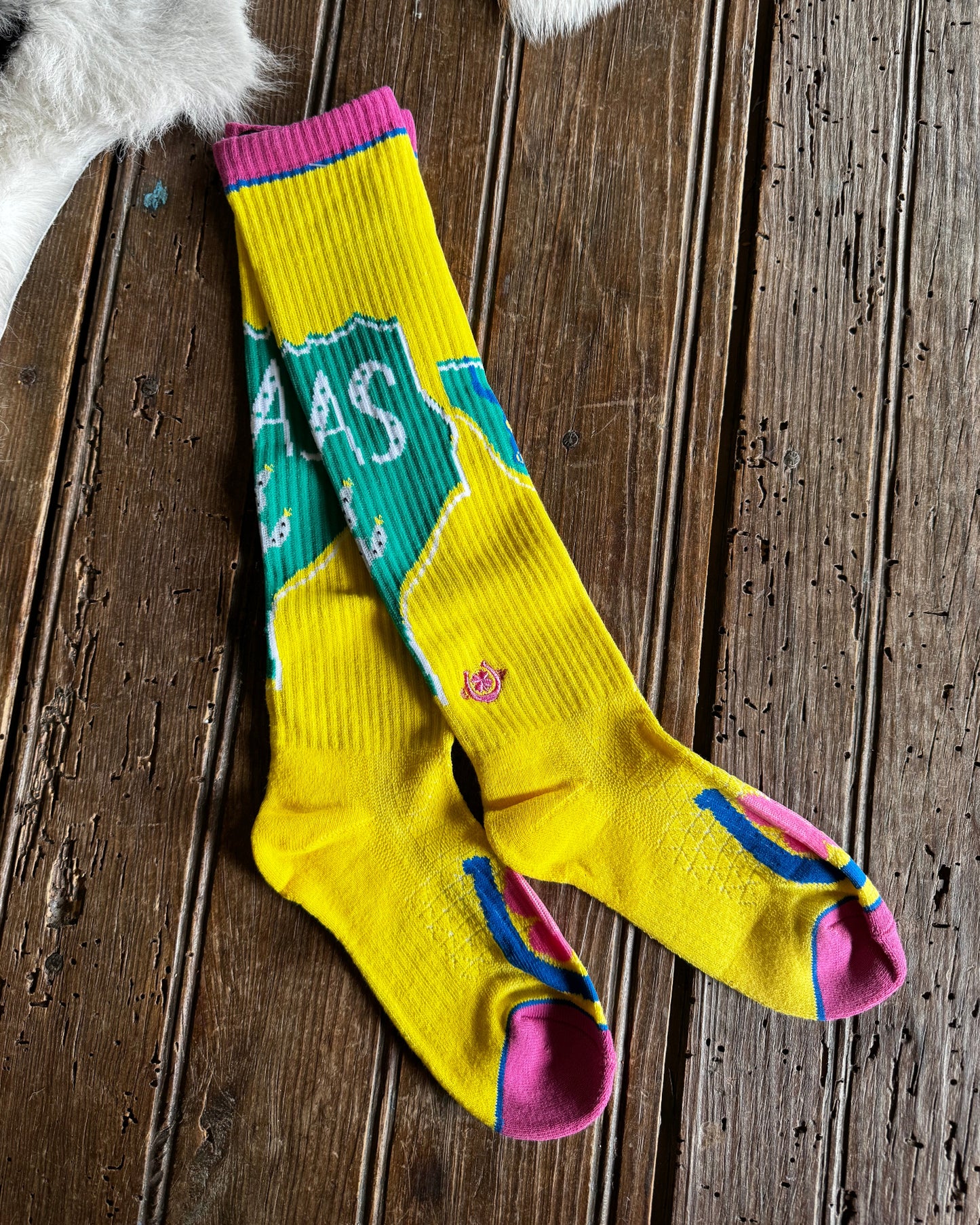 A pair of bright yellow crew socks with pink, teal, and blue graphic details, laid flat on a rustic dark wood plank surface.