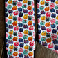 A close-up of a white fabric with a colorful repeating pattern of cartoon pig-like creatures in pink, orange, yellow, teal, and purple, laid flat on a rustic wooden surface.