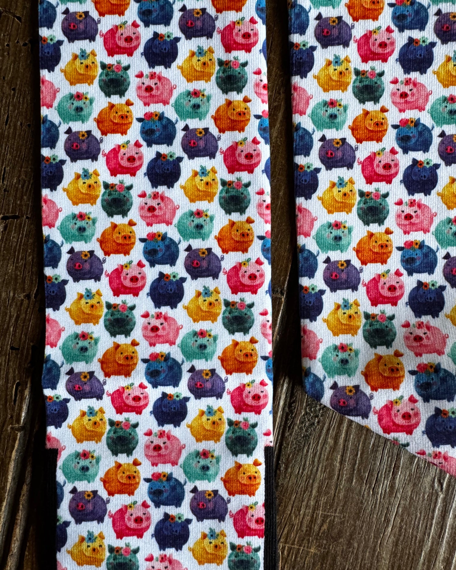 A close-up of a white fabric with a colorful repeating pattern of cartoon pig-like creatures in pink, orange, yellow, teal, and purple, laid flat on a rustic wooden surface.