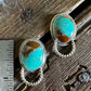 A pair of silver-toned drop earrings with oval turquoise stones and twisted rope details, displayed on a rustic wooden surface next to a metal ruler for scale.