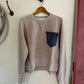 Beige sweatshirt with a blue pocket on a hanger against a white wall.
