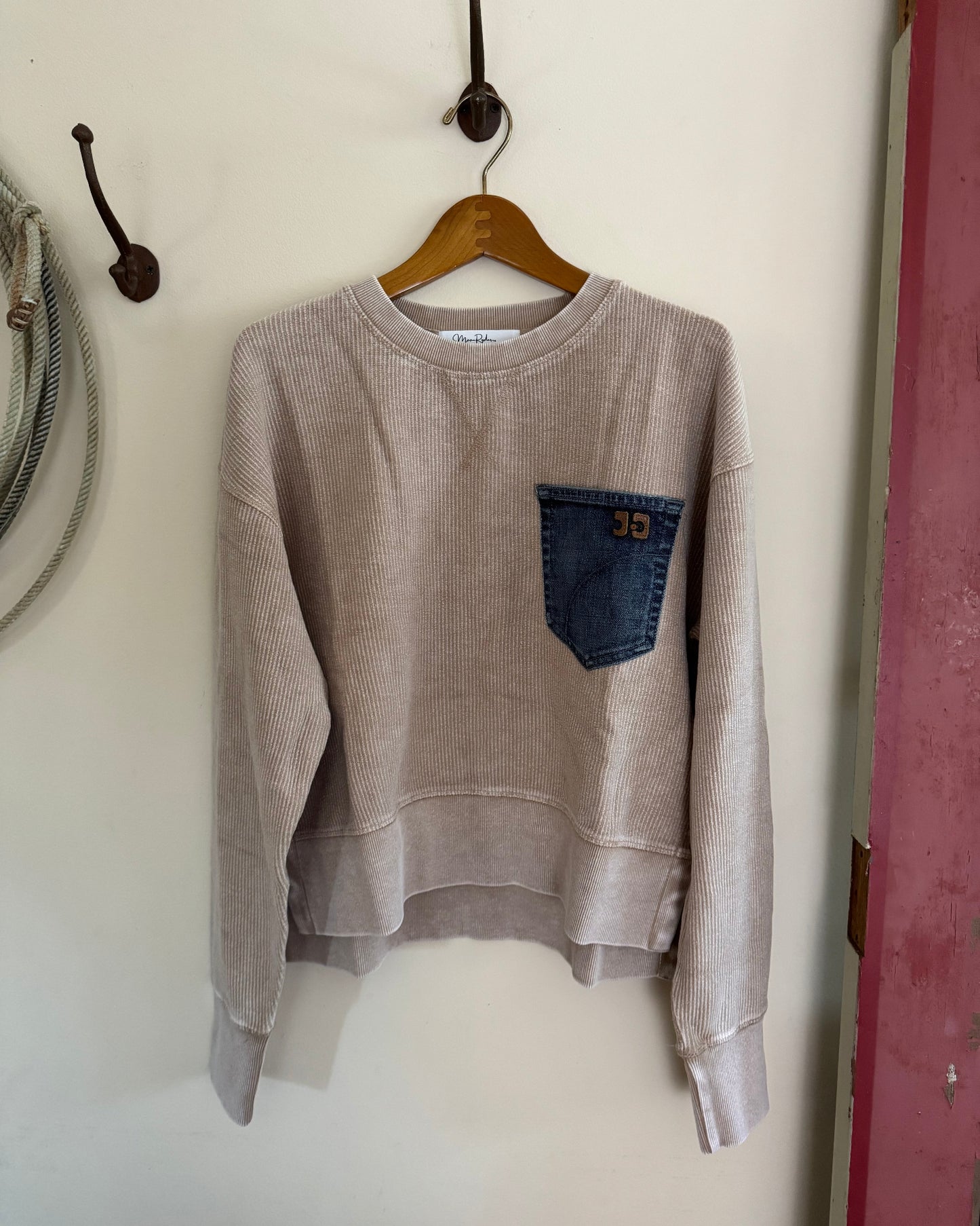 Beige sweatshirt with a blue pocket on a hanger against a white wall.