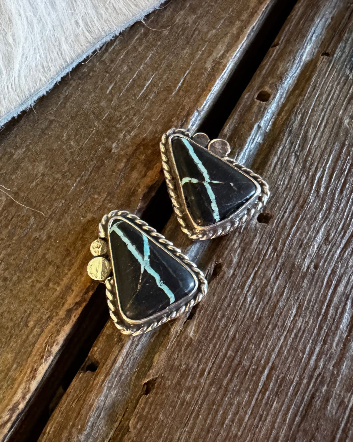 A pair of triangular black stone earrings with turquoise veining and gold-toned bezel settings, resting on a rustic wooden surface next to a white faux fur trim.