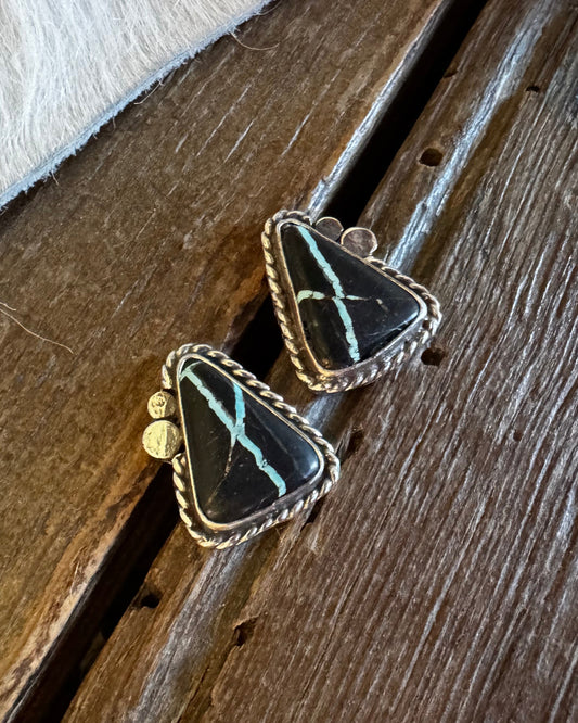 A pair of triangular black stone earrings with turquoise veining and gold-toned bezel settings, resting on a rustic wooden surface next to a white faux fur trim.