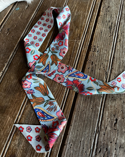 A long scarf with a colorful floral and paisley pattern on a light blue base, laid out on a rustic wooden plank surface.