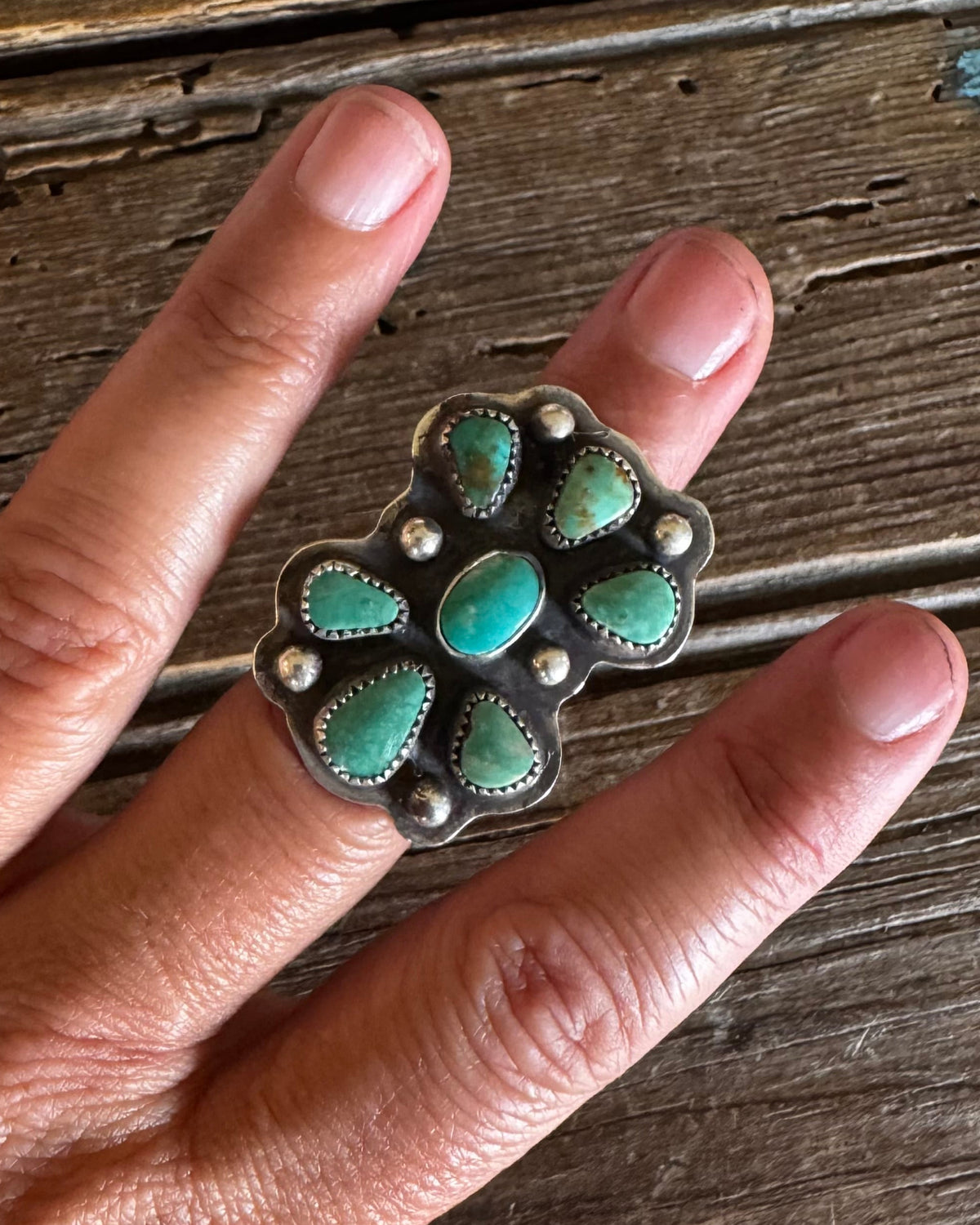 A close-up of a hand wearing a large silver ring set with multiple oval turquoise stones, resting on a rustic wooden surface.