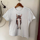 A white short-sleeve t-shirt hanging on a wooden hanger against a beige wall, featuring a graphic print of a horse head wearing a lasso.