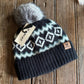 Knitted beanie with pattern and pom-pom on a wooden surface, featuring a brand tag.