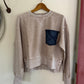 Beige sweatshirt with a denim pocket hanging on a wooden hanger against a white wall.