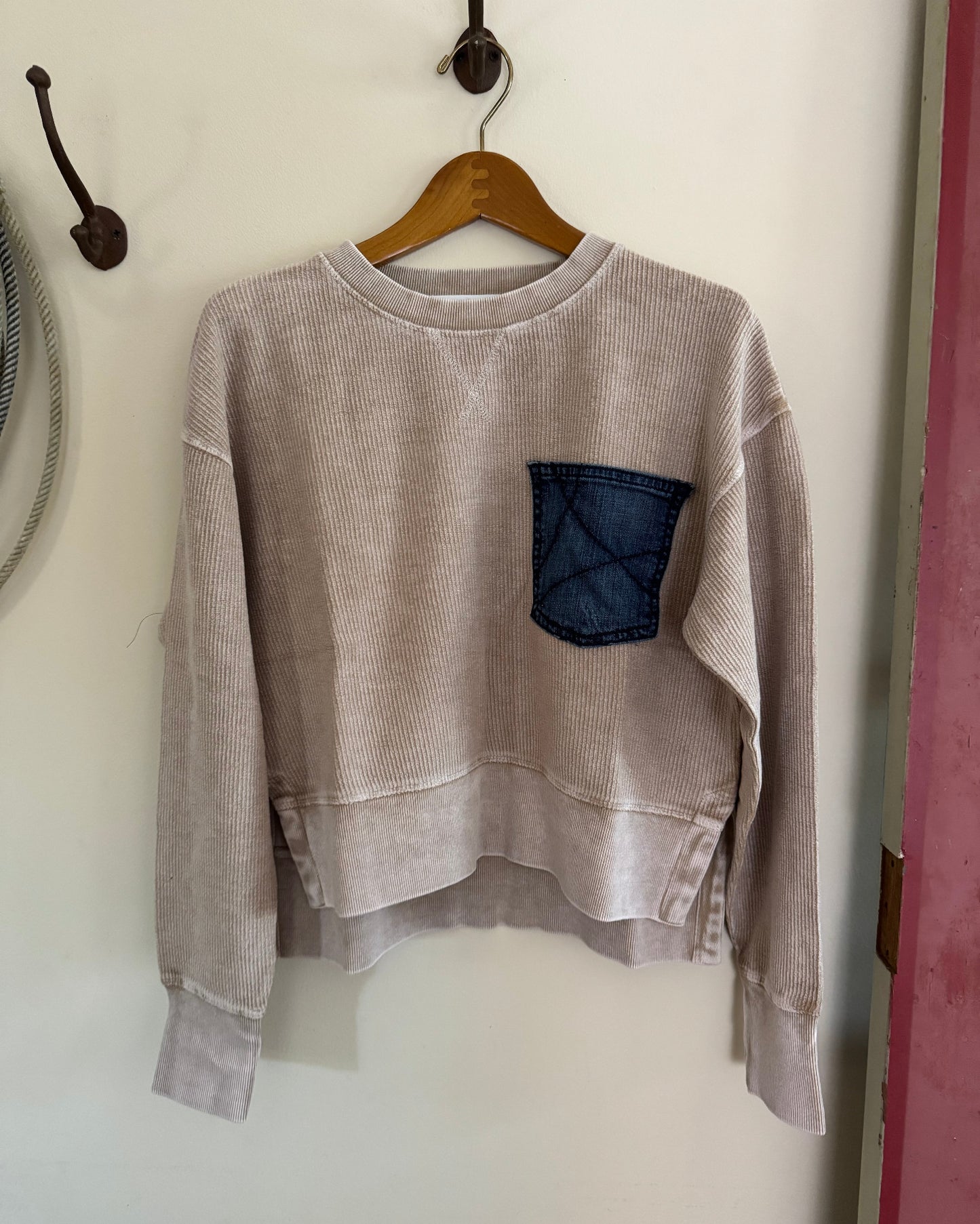 Beige sweatshirt with a denim pocket hanging on a wooden hanger against a white wall.