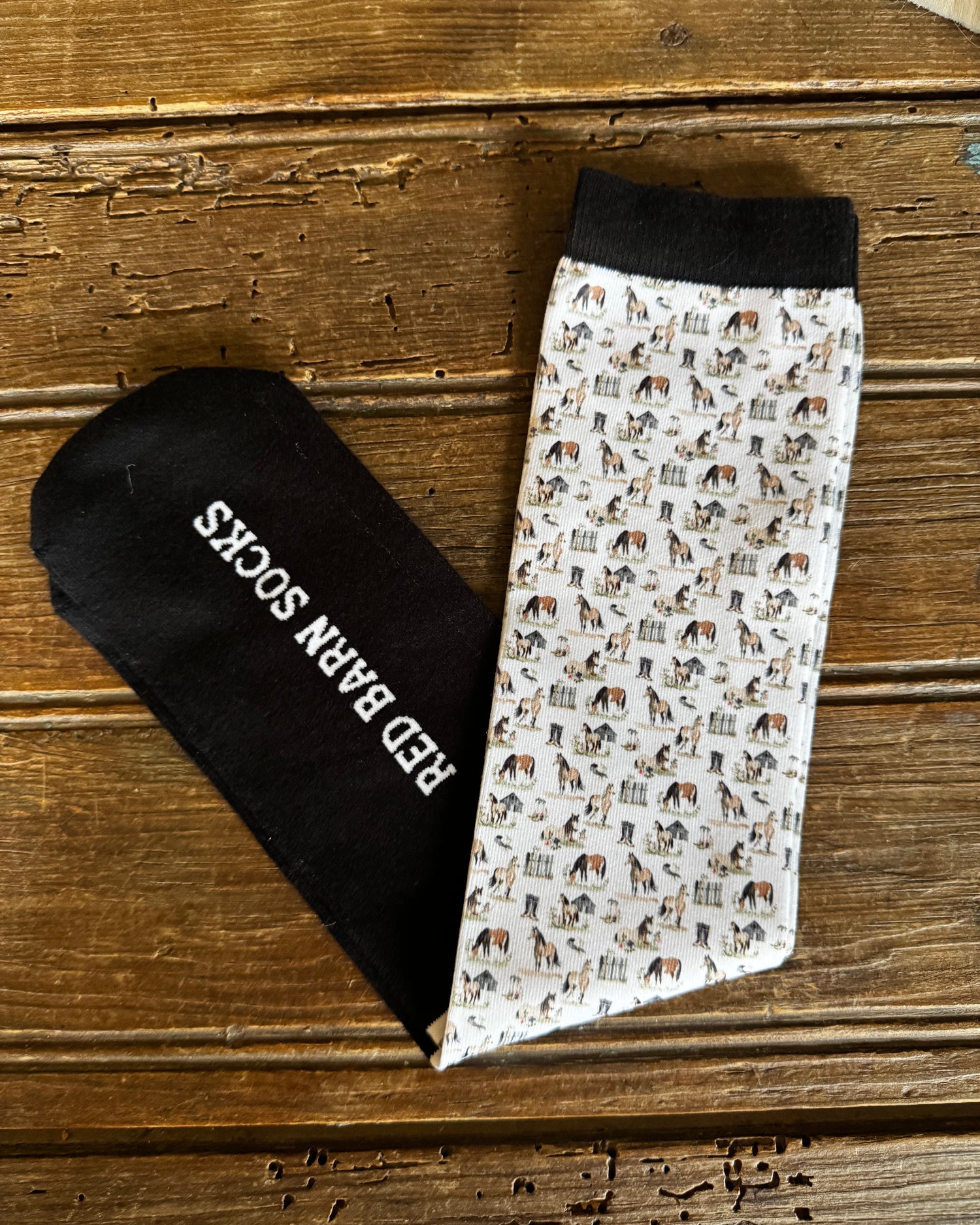 Pair of socks with horse pattern and black sock with 'Red Barn Socks' text on a wooden surface