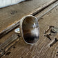 A silver ring with a large, polished oval cabochon gemstone featuring brown, black, and white marbled patterns, resting on a rustic wooden surface with a white fur trim at the top.