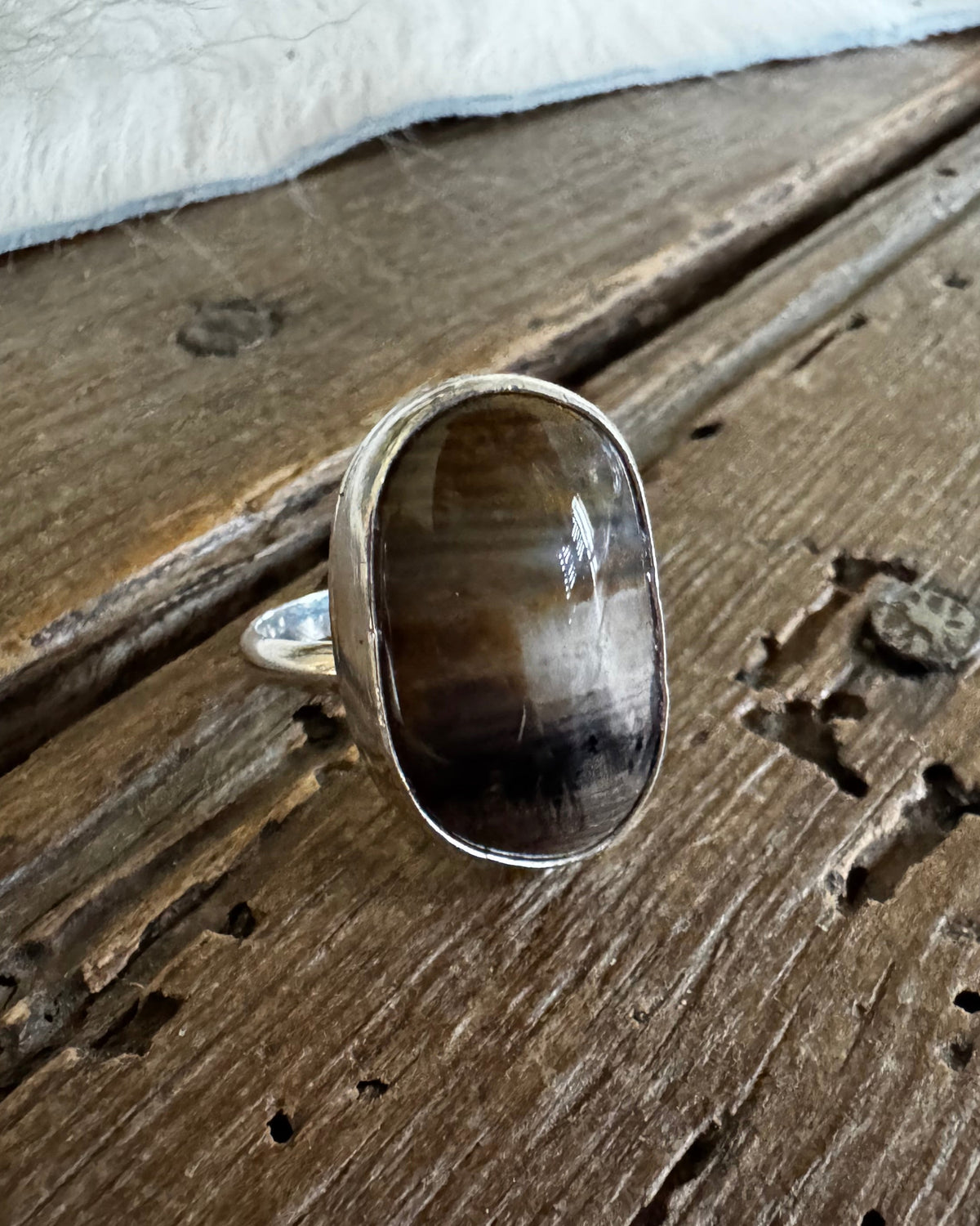 A silver ring with a large, polished oval cabochon gemstone featuring brown, black, and white marbled patterns, resting on a rustic wooden surface with a white fur trim at the top.