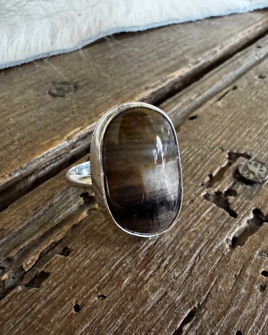 A silver ring with a large, polished oval cabochon gemstone featuring brown, black, and white marbled patterns, resting on a rustic wooden surface with a white fur trim at the top.