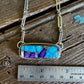 Necklace with a rectangular pendant featuring blue and purple stones, displayed on a wooden surface with a ruler for scale.
