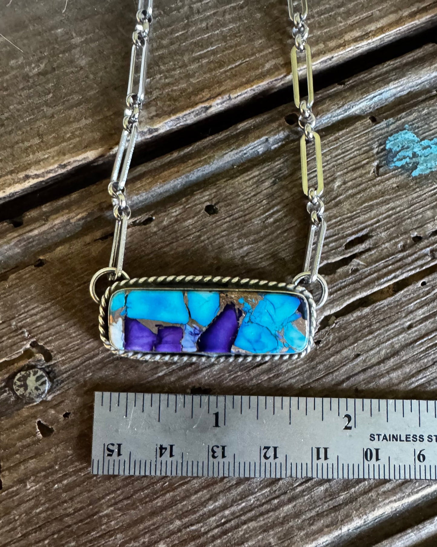 Necklace with a rectangular pendant featuring blue and purple stones, displayed on a wooden surface with a ruler for scale.