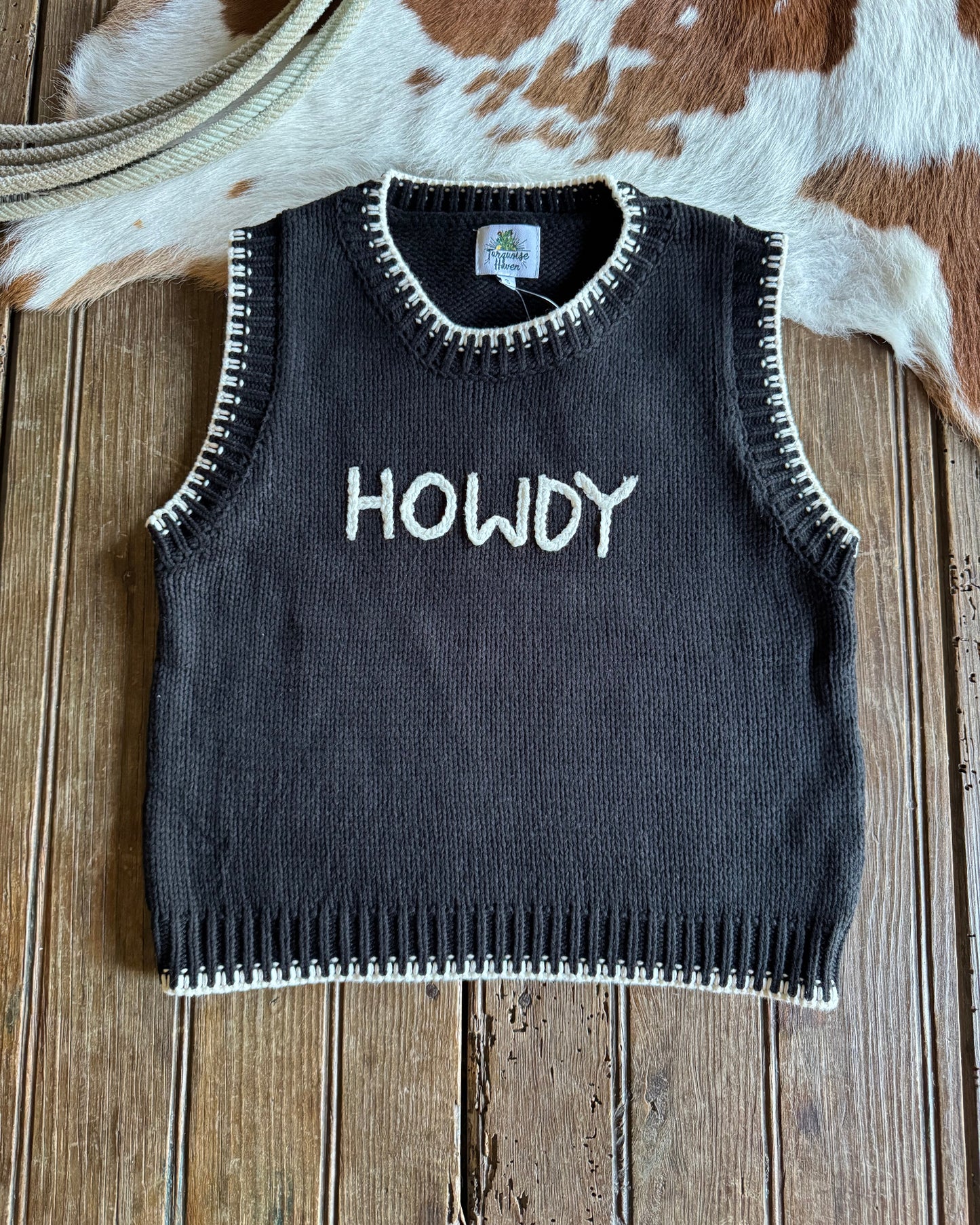 Black sweater with 'HOWDY' text on a wooden surface next to a cow.