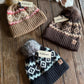 Three knit hats with patterned designs and pom-poms on a wooden surface, Ariat branded.