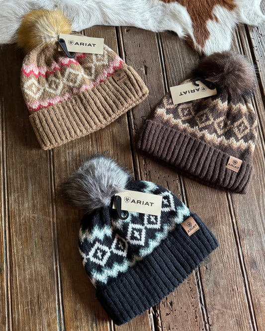 Three knit hats with patterned designs and pom-poms on a wooden surface, Ariat branded.