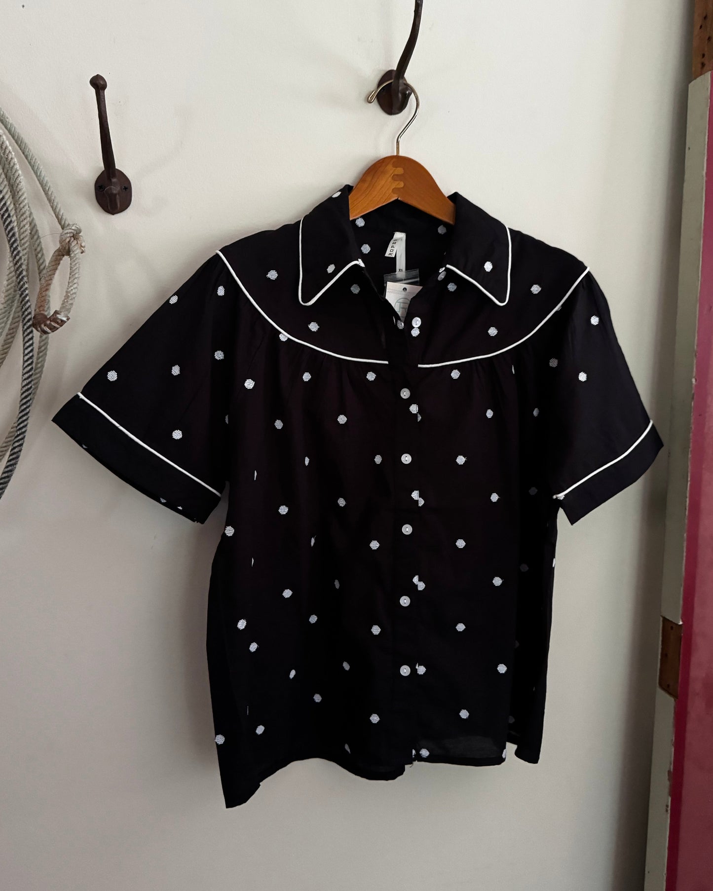 Black blouse with white polka dots on a wooden hanger against a white wall.