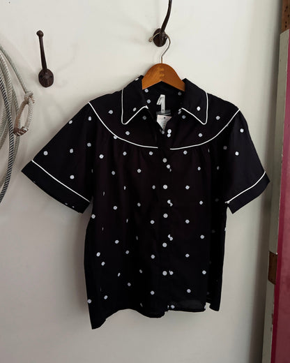 Black blouse with white polka dots on a wooden hanger against a white wall.