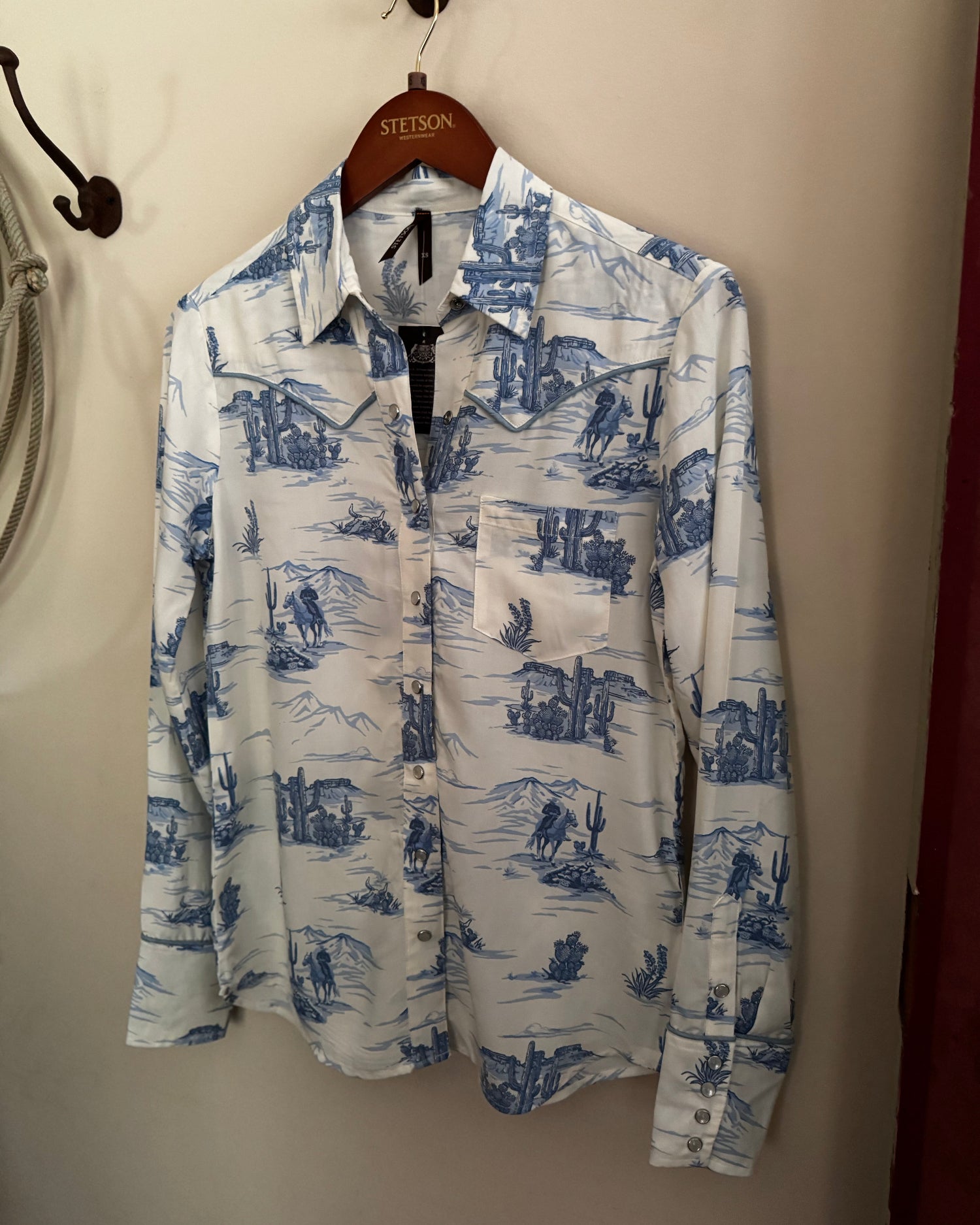 White shirt with blue desert landscape print hanging on a hanger against a beige wall.