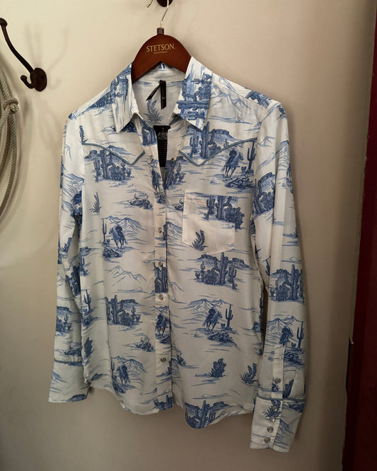 White shirt with blue desert landscape print hanging on a hanger against a beige wall.