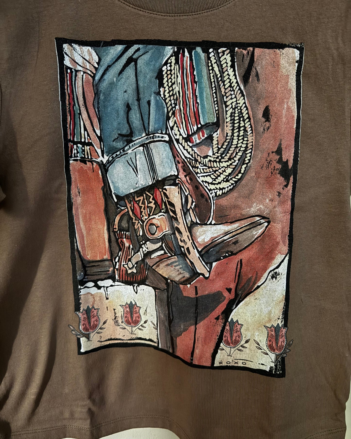 A brown short-sleeve graphic t-shirt featuring a large rectangular print of a cowboy boot, lasso, and western clothing in earthy tones of red, blue, beige, and brown.