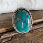 A silver-toned ring with an oval turquoise stone, resting on a rustic wooden surface with white faux fur in the background.