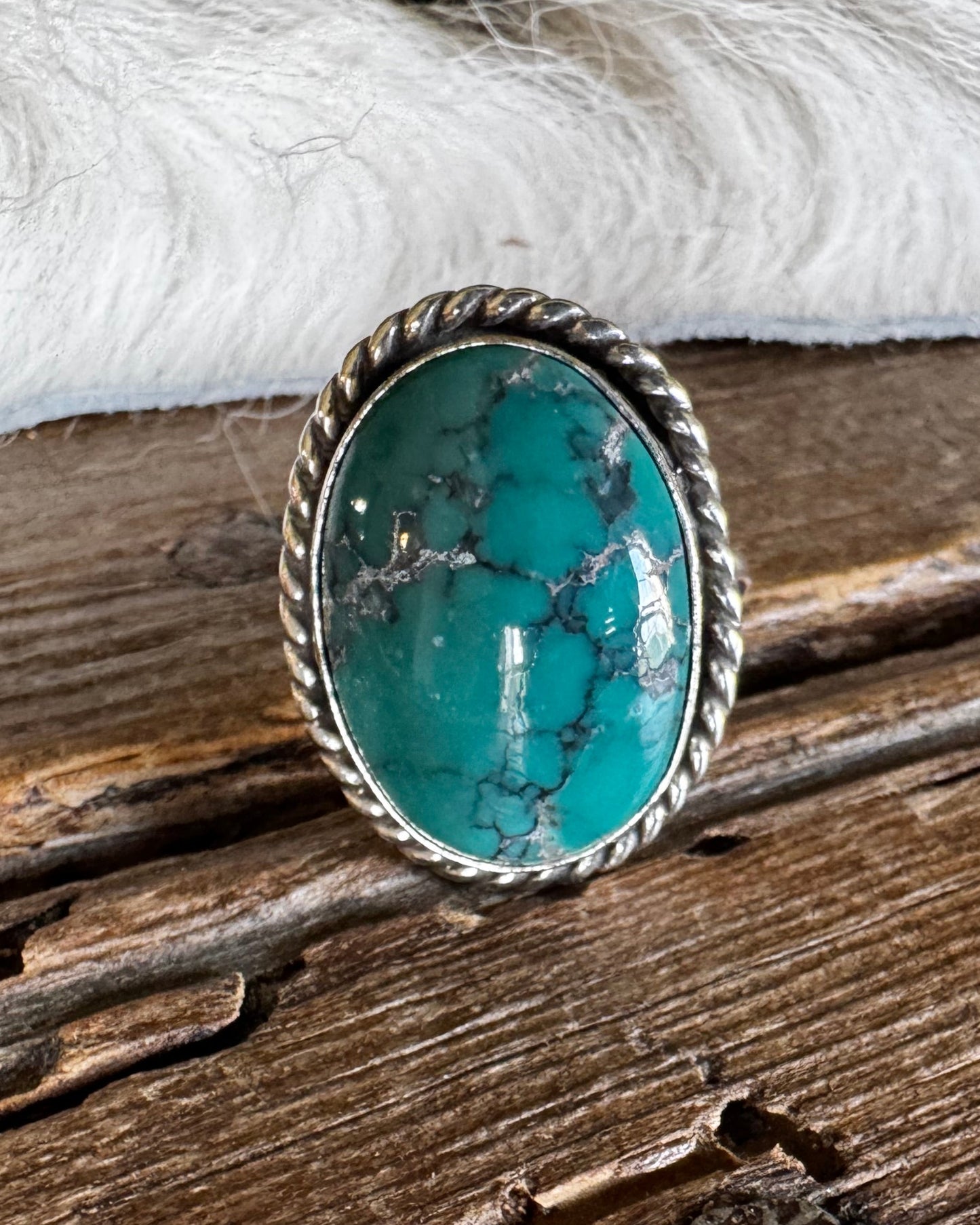 A silver-toned ring with an oval turquoise stone, resting on a rustic wooden surface with white faux fur in the background.