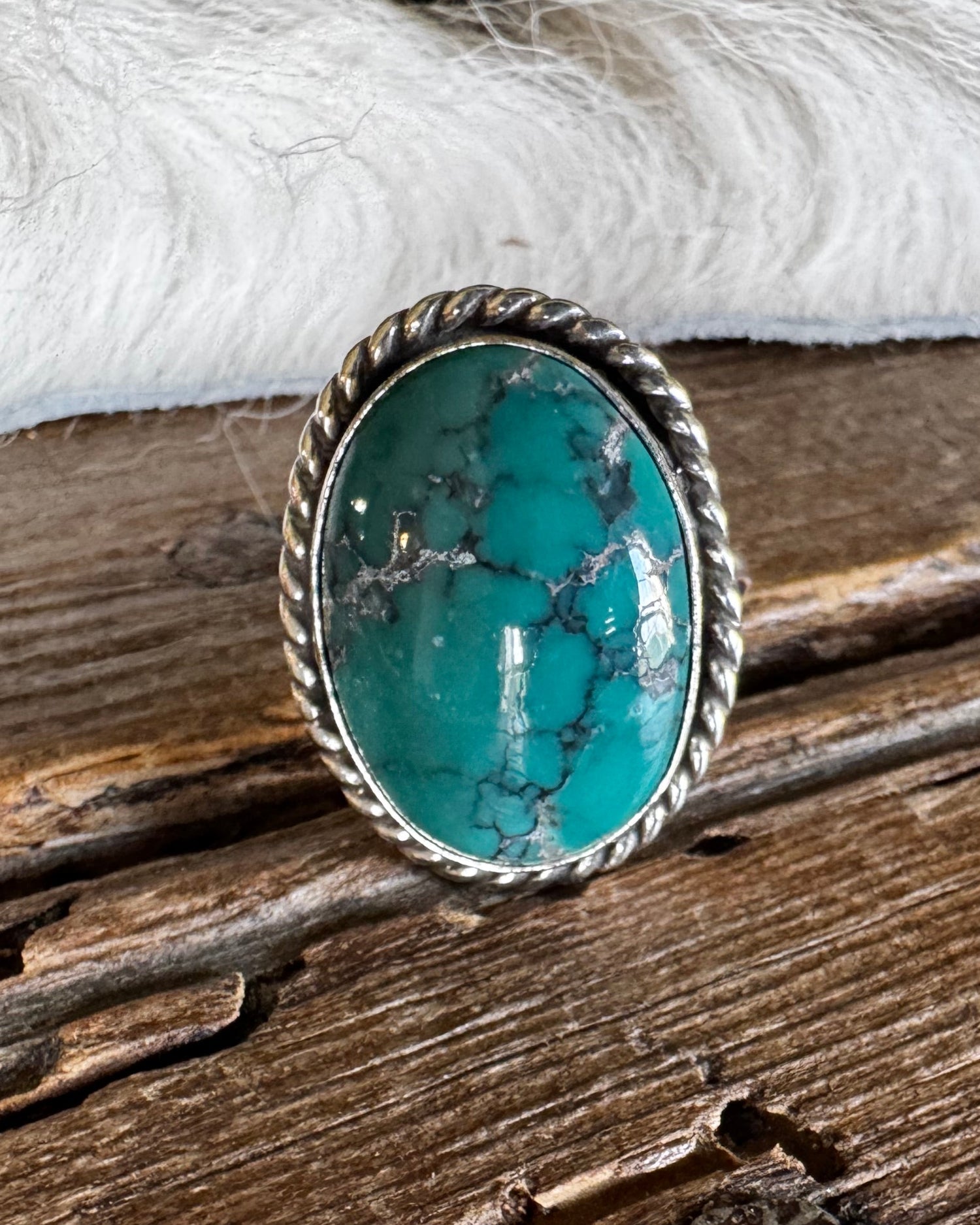 A silver-toned ring with an oval turquoise stone, resting on a rustic wooden surface with white faux fur in the background.