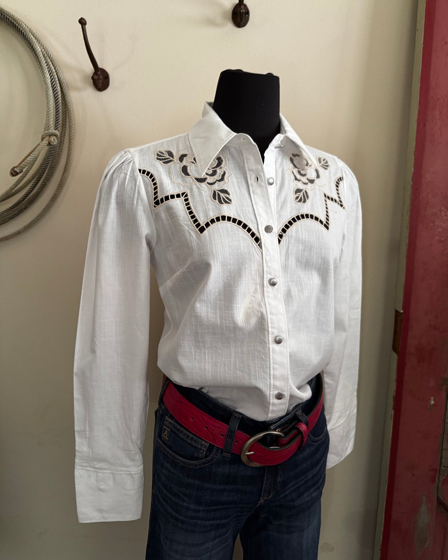 White embroidered shirt on a mannequin with a red belt against a neutral wall.