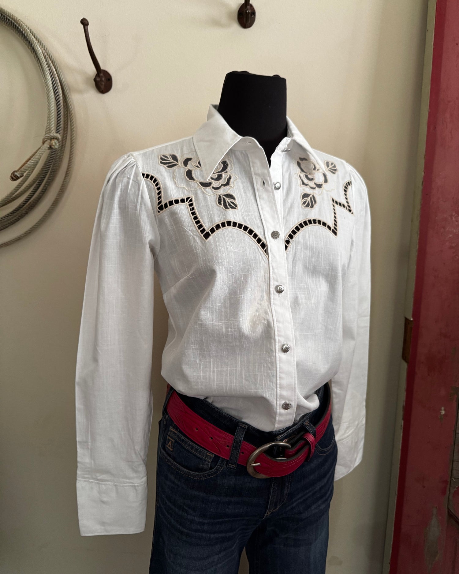 White embroidered shirt on a mannequin with a red belt against a neutral wall.