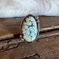 A Southwestern-style sterling silver ring featuring an oval turquoise stone with prominent black matrix veining, resting on a rustic wooden surface with white fur in the background.