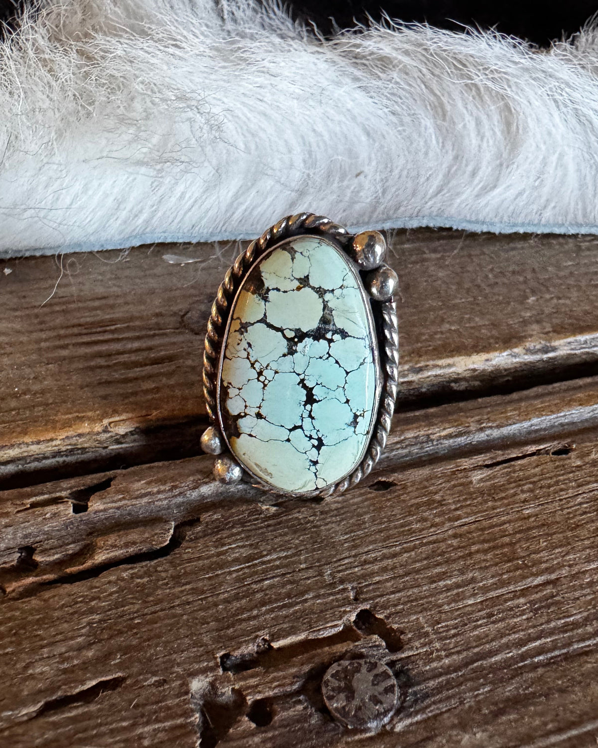 A Southwestern-style sterling silver ring featuring an oval turquoise stone with prominent black matrix veining, resting on a rustic wooden surface with white fur in the background.