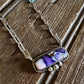 A silver-toned chain necklace with a rectangular pendant featuring purple, white, and pink agate stone, displayed on a rustic wooden surface with a white faux fur accent.