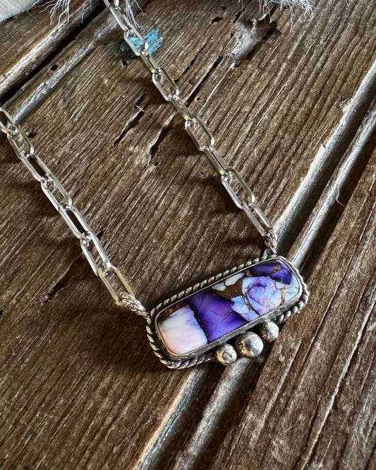 A silver-toned chain necklace with a rectangular pendant featuring purple, white, and pink agate stone, displayed on a rustic wooden surface with a white faux fur accent.