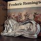 A polished silver horse-shaped serving tray or dish displayed on a dark wooden surface in front of a vintage book titled 'Frederic Remington'.