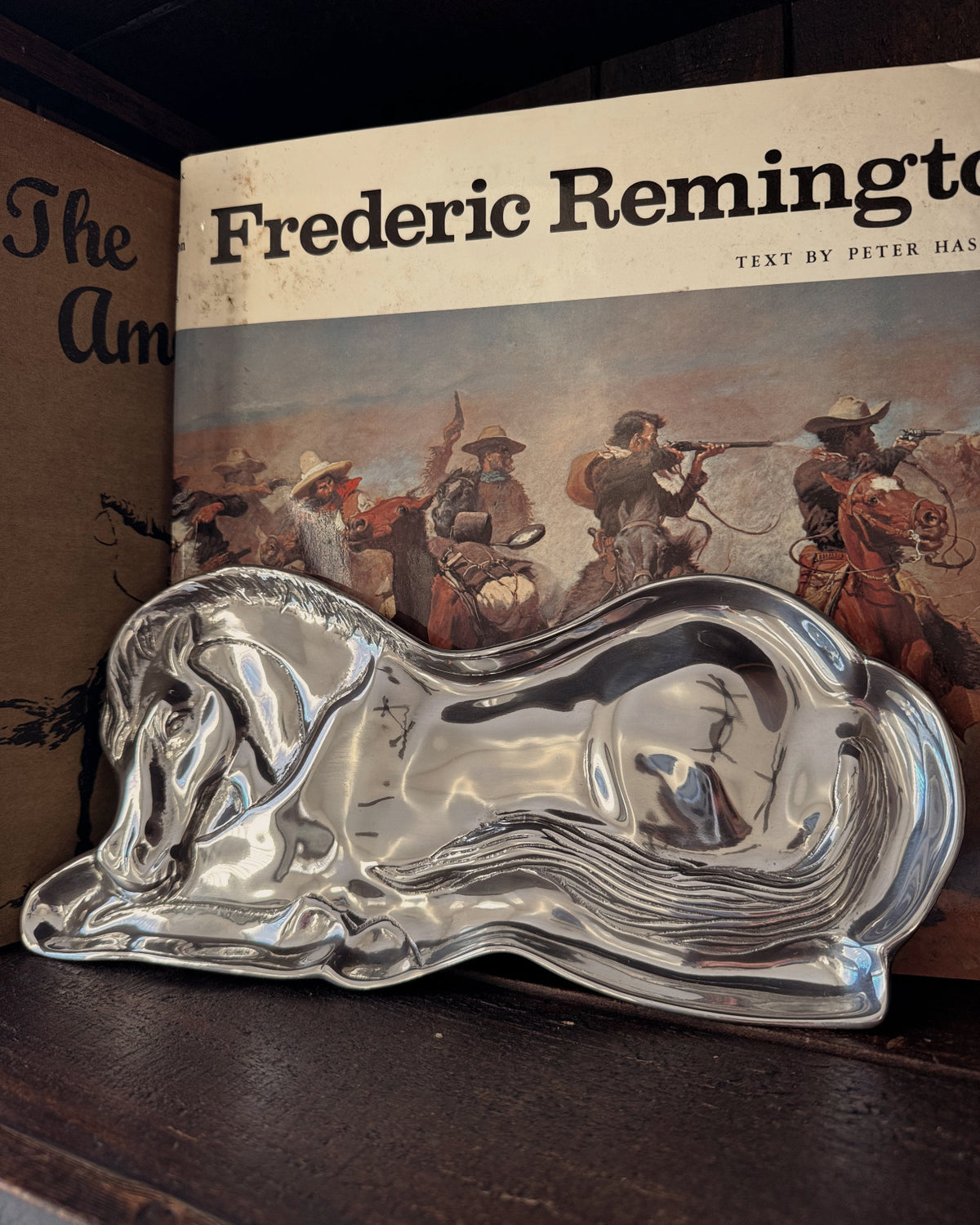 A polished silver horse-shaped serving tray or dish displayed on a dark wooden surface in front of a vintage book titled 'Frederic Remington'.