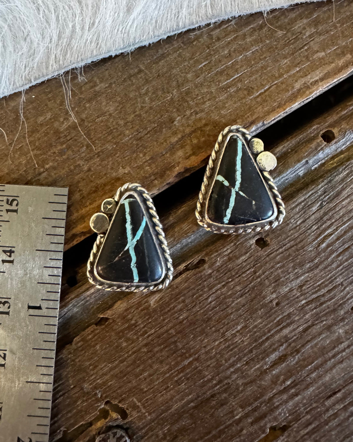 A pair of triangular turquoise and black stone stud earrings with silver-toned bezel settings, displayed on a rustic wooden surface next to a metal ruler for scale.