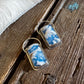 A pair of gold-tone rectangular earrings with blue and white cloud-patterned resin inlays, displayed on a rustic wooden surface next to a white faux-fur trim.