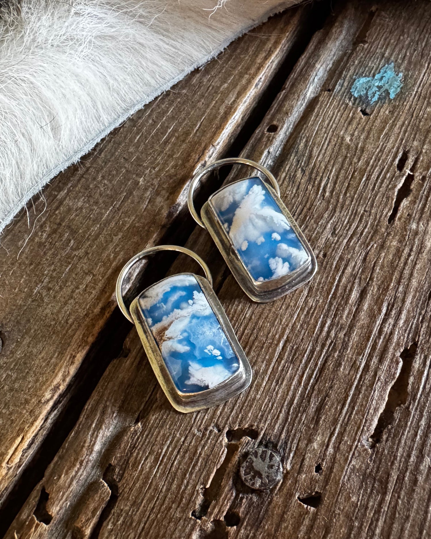 A pair of gold-tone rectangular earrings with blue and white cloud-patterned resin inlays, displayed on a rustic wooden surface next to a white faux-fur trim.
