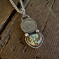 A silver-toned pendant necklace featuring a round coin-style charm and a larger heart-shaped stone pendant with turquoise-like blue and brown marbling, displayed on a rustic dark wood surface.